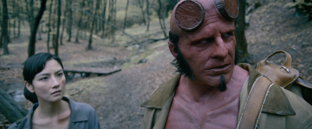 Agent Bobbie Jo Song (Adeline Rudolph) and Hellboy (Jack Kesy) stand on a forest path together, looking wary, in Hellboy: The Crooked Man.