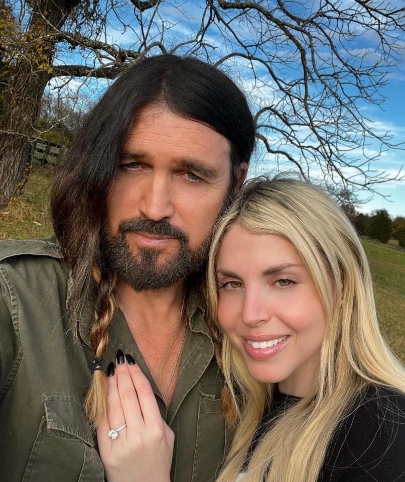 Billy Ray Cyrus and Firerose embracing outdoors, she showing off her engagement ring.