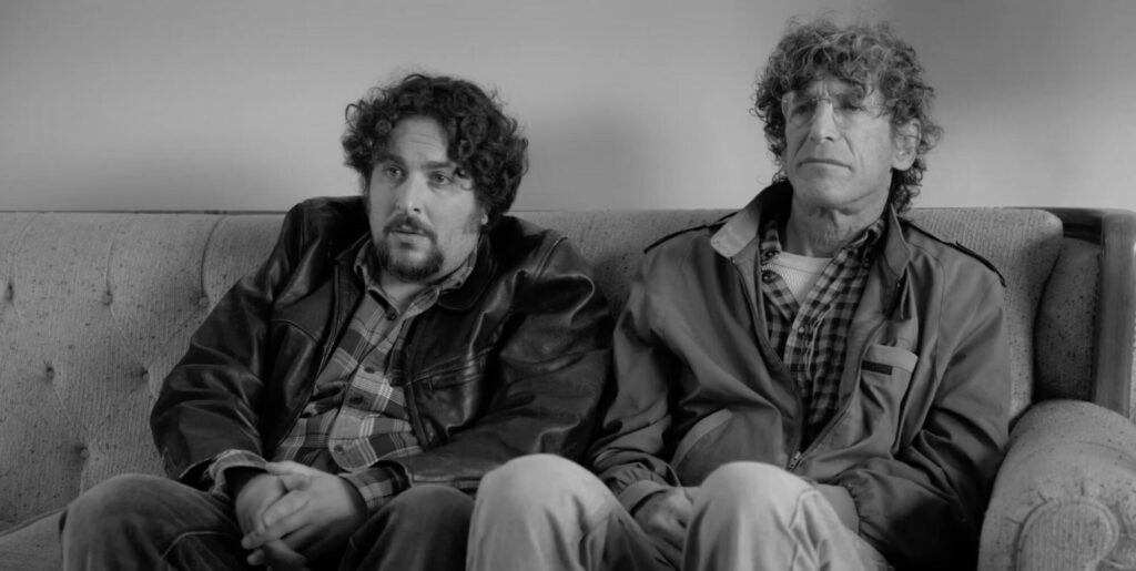 Two men sit uncomfortably next to each other on a couch.