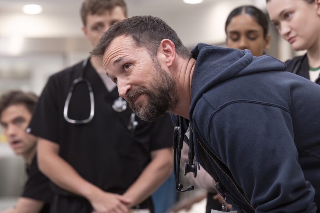 Noah Wyle, surrounded by student doctors, looks up from a hunched position in The Pitt