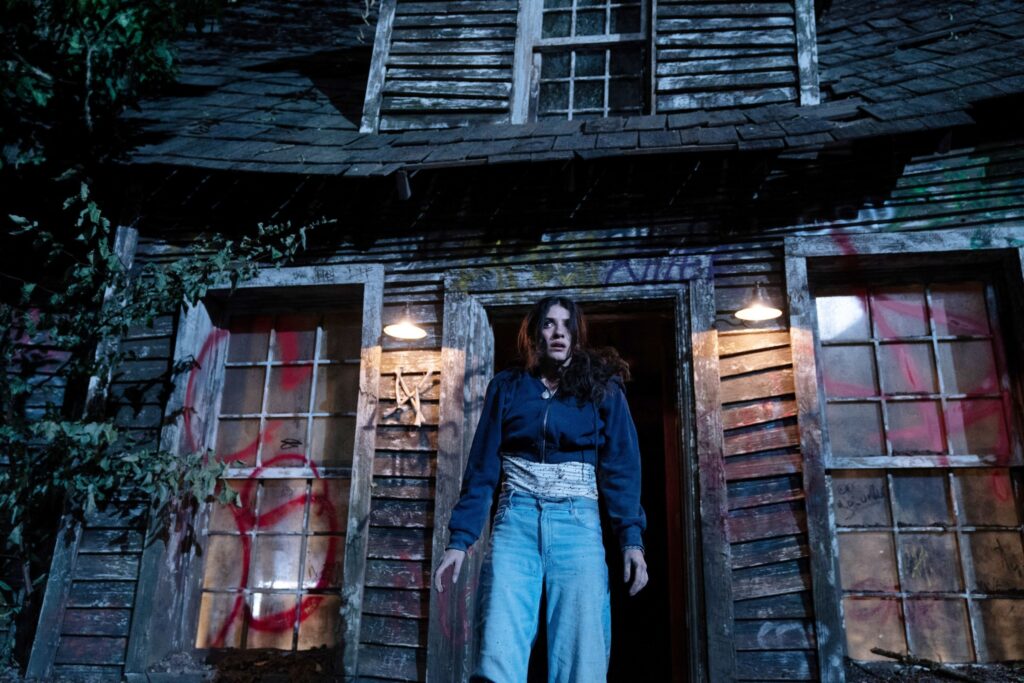 Clover (Ella Rubin), a teenager in a high-waisted dark blue sweater and light blue pants, stands in the front doorway of a battered-looking house covered with red spray-paint graffiti in Until Dawn