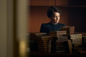 Keeley Hawes, in a black dress, sits at a table behind piles of leather-bound books in "Miss Austen."