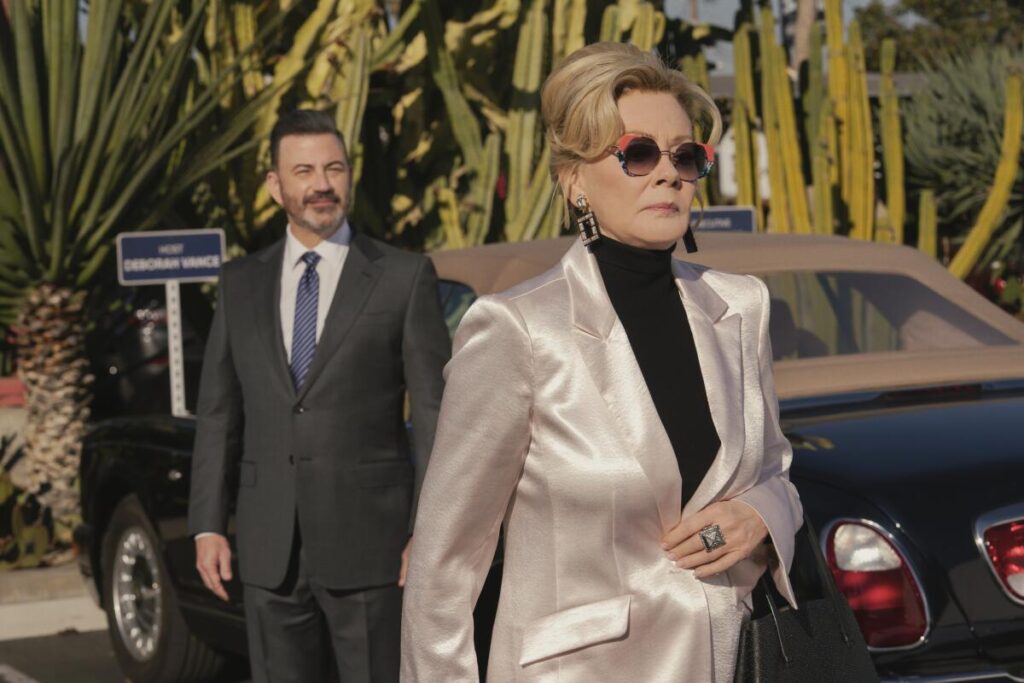 A man in a suit and blue tie looks at the back of a woman in a white blazer and black turtleneck as she walks away.
