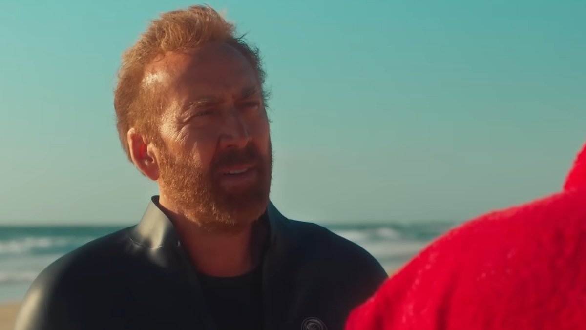 Nic Cage on the beach in a wetsuit speaks to a man in red in The Surfer