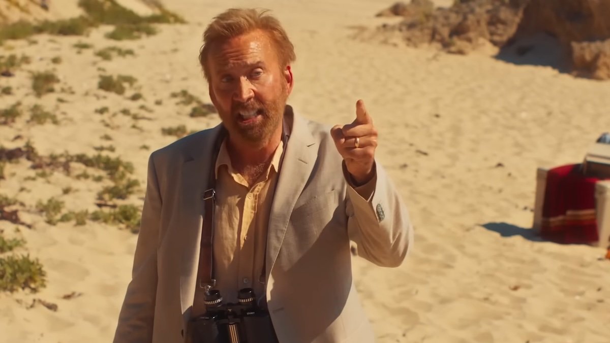 Nic Cage in a suit on sand pointing up in The Surfer