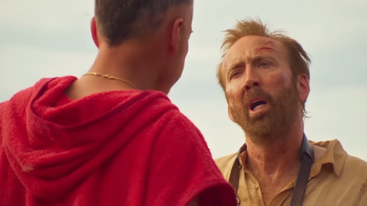 A blooded Nic Cage looking confused peaks to a man in red in The Surfer