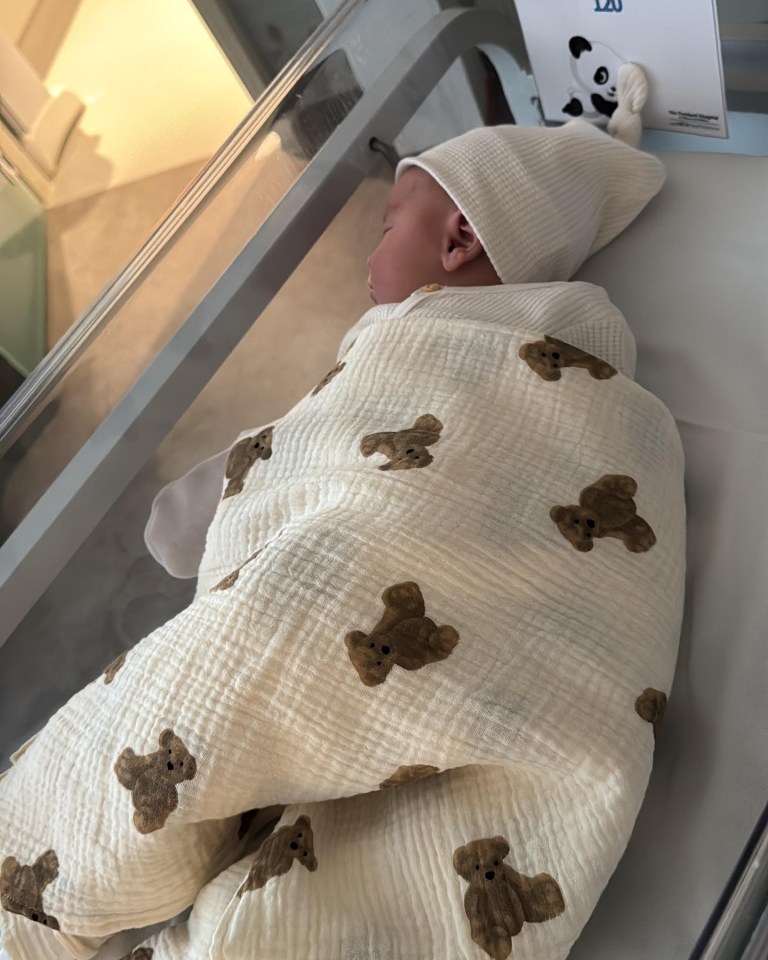 Newborn baby sleeping in a hospital bassinet wrapped in a teddy bear patterned blanket.