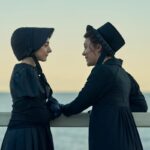 Two women in black bonnets and dresses stand and look at each near a railing overlooking a body of water.