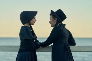 Two women in black bonnets and dresses stand and look at each near a railing overlooking a body of water.