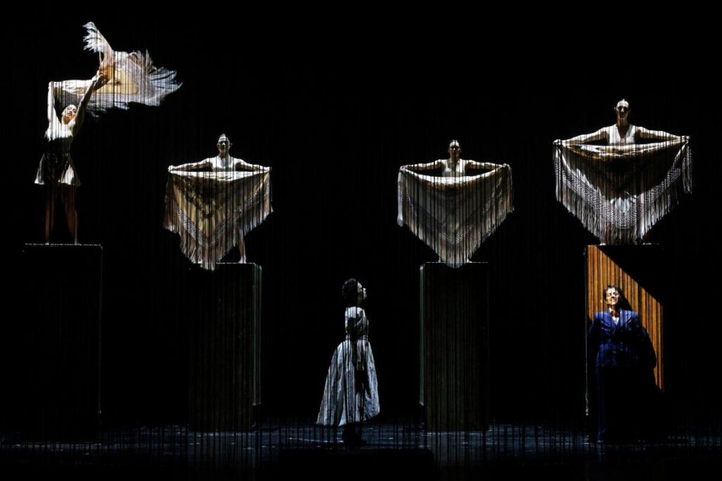 Two characters sing onstage as ghostly figures seem to hover above.