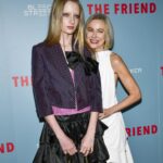 Kai Schreiber in a dark outfit and Naomi Watts in a white one stand in front of a backdrop
