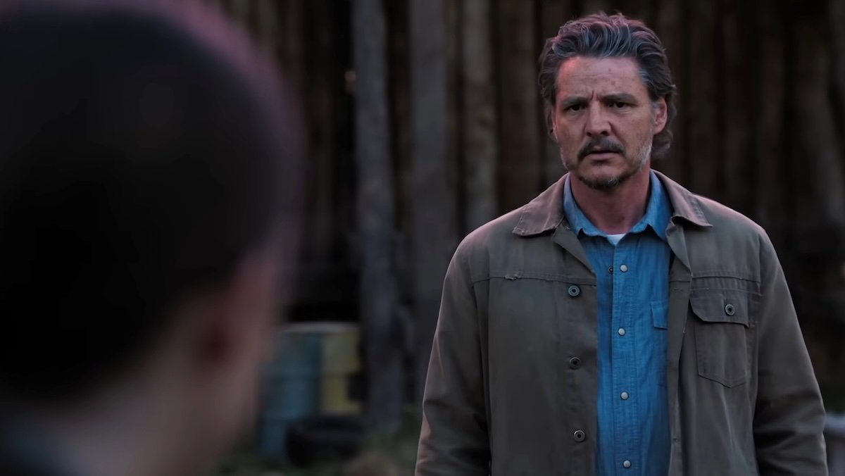 Pedro Pascal as Joel stands outside in a brown coat and blue shirt on The Last of Us