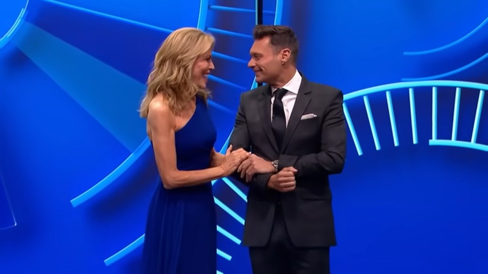 Two people in formal wear stand in front of a blue Wheel of Fortune-like set.