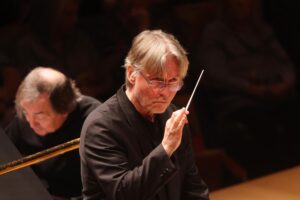 Esa-Pekka Salonen holds his baton while conducting the L.A. Phil, with pianist Pierre-Laurent Aimard seated behind him.