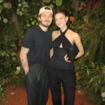 Brooklyn Beckham and Nicola Peltz Beckham posing together in front of a floral wall.