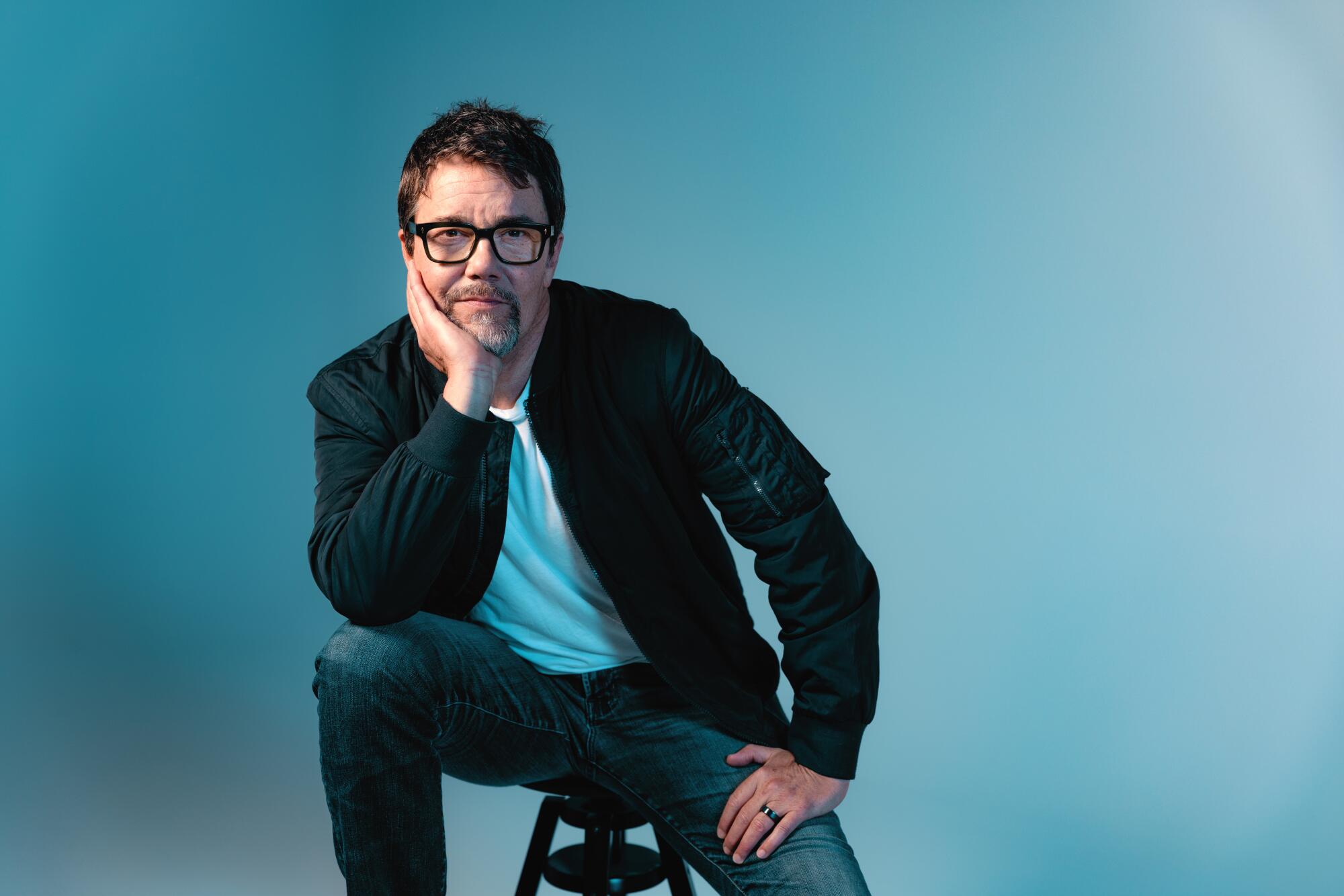 A man in black-rimmed glasses, black jacket and jeans sits on a stool with his arm on his knee and head resting on his hand.