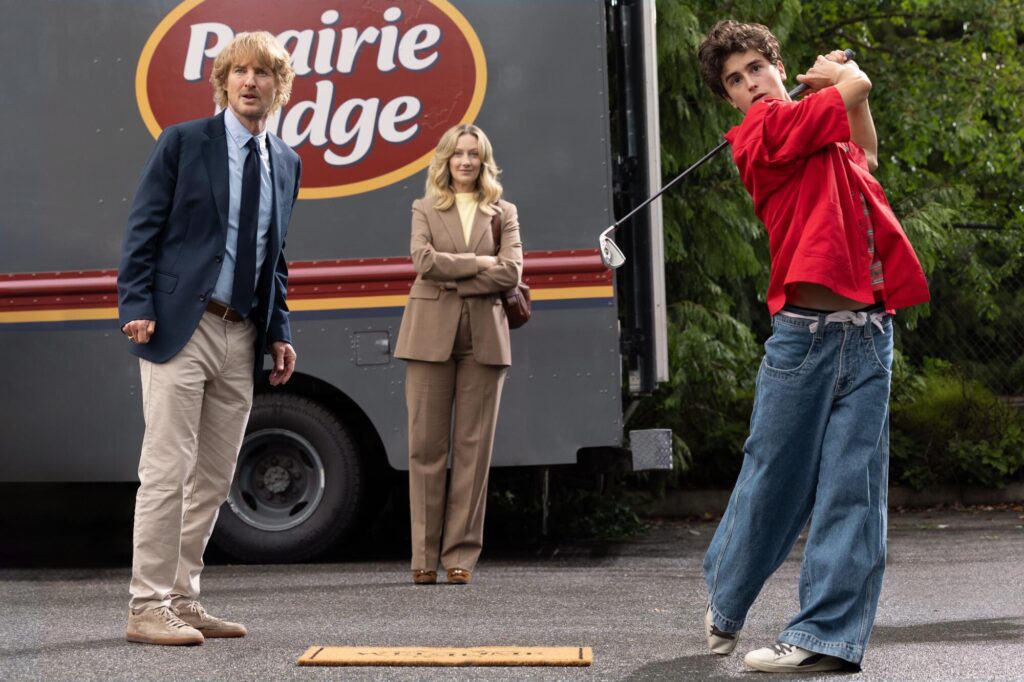 Two older adults stand near a box truck as a teenage boy in a red shirt and wide-leg jeans swings a golf club.