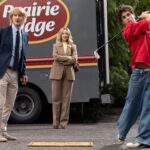 Two older adults stand near a box truck as a teenage boy in a red shirt and wide-leg jeans swings a golf club.