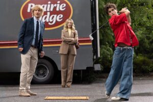 Two older adults stand near a box truck as a teenage boy in a red shirt and wide-leg jeans swings a golf club.