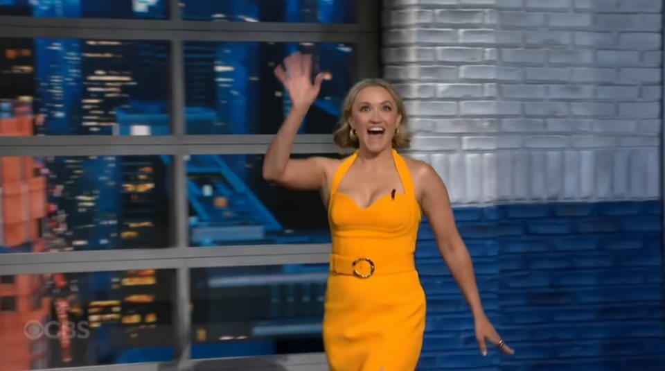 Emily Osment waving in a yellow dress.