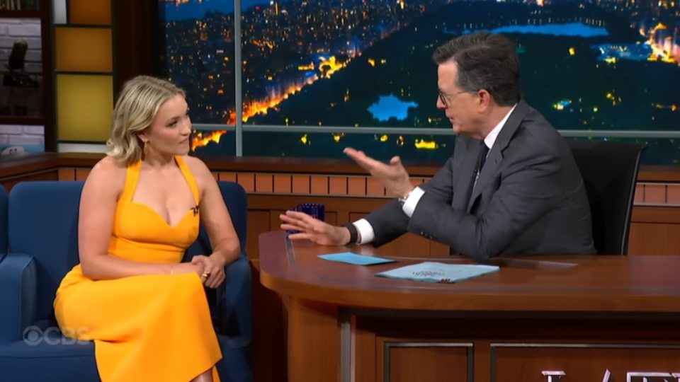 Emily Osment in a yellow dress being interviewed on a late-night talk show.