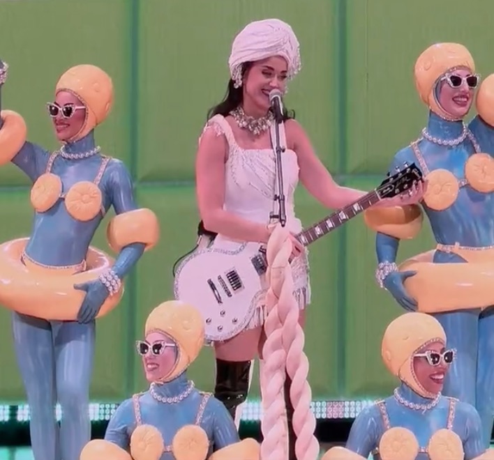 Katy Perry playing guitar next to dangers in skintight blue suits with peach colored hats, bras, inner tubes around their bottoms and arms along with large white pearl accents