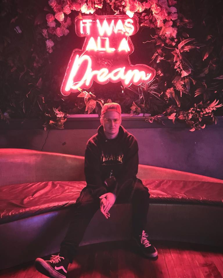 Adam Ramey, lead singer of Dropout Kings, sitting on a curved bench in front of a neon sign that says "It Was All A Dream".