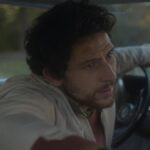 A man in a white jacket leans forward in a car nervously.