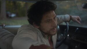 A man in a white jacket leans forward in a car nervously.