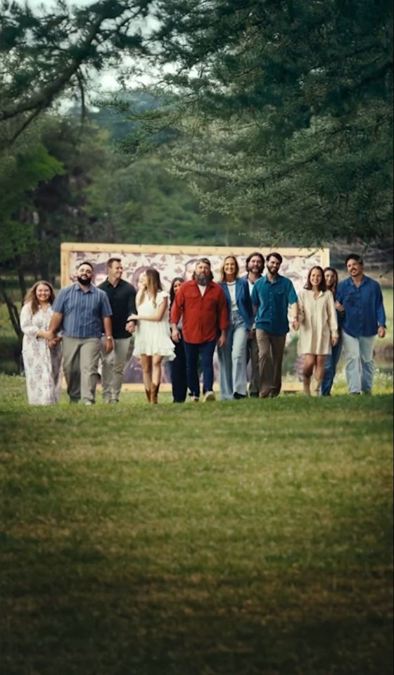 Large group of people walking on a grassy field in front of a large painting.