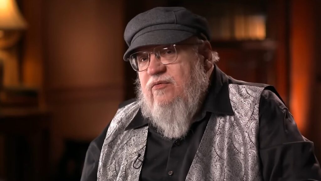 George R.R. Martin in his hat and gray vest sitting for an interview