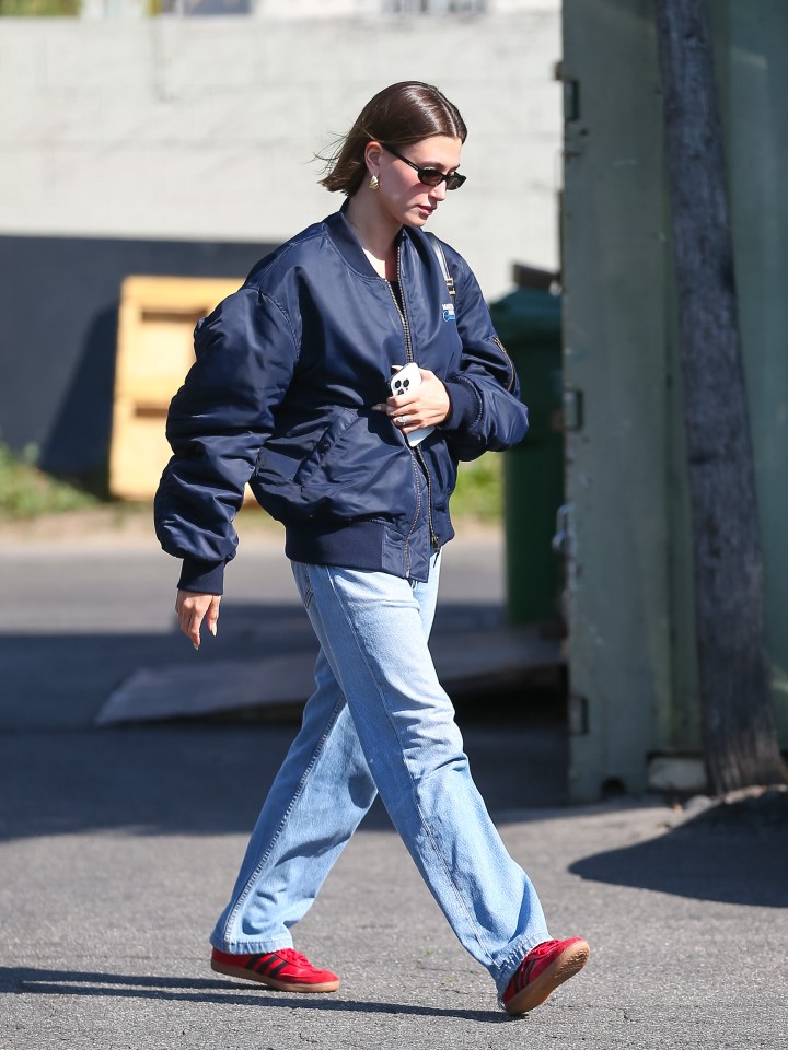 Hailey Bieber walking down the street in a navy bomber jacket, light blue jeans, and red Adidas sneakers.