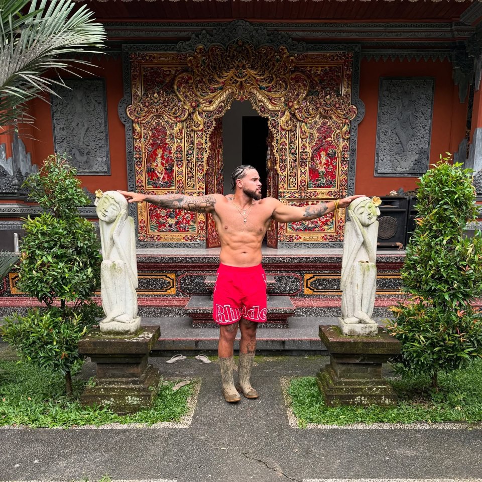 Shirtless man in red shorts standing between two statues in front of an ornate door.