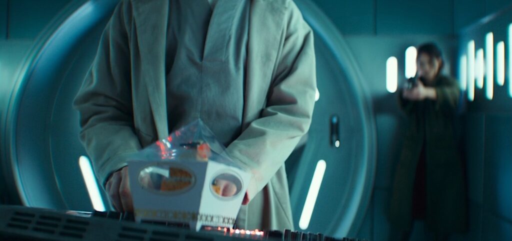 A shot from season 2, episode 6 of Andor, with Bix (Adria Arjona) in the background, out of focus, pointing a blaster at Imperial torturer Doctor Gorst (Joshua James), whose back is to her. He’s standing in the foreground at a computer panel, having just put down a fancy cupcake in a decorative box on the panel in front of him, in the extreme foreground.