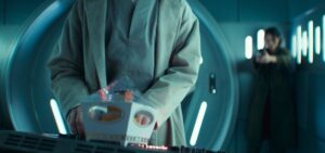 A shot from season 2, episode 6 of Andor, with Bix (Adria Arjona) in the background, out of focus, pointing a blaster at Imperial torturer Doctor Gorst (Joshua James), whose back is to her. He’s standing in the foreground at a computer panel, having just put down a fancy cupcake in a decorative box on the panel in front of him, in the extreme foreground.
