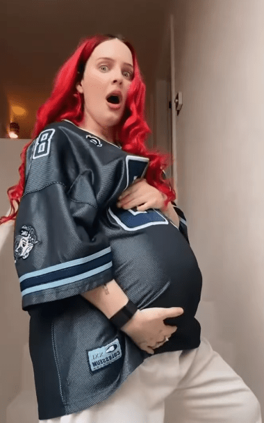 Pregnant woman with red hair wearing a jersey, looking surprised.