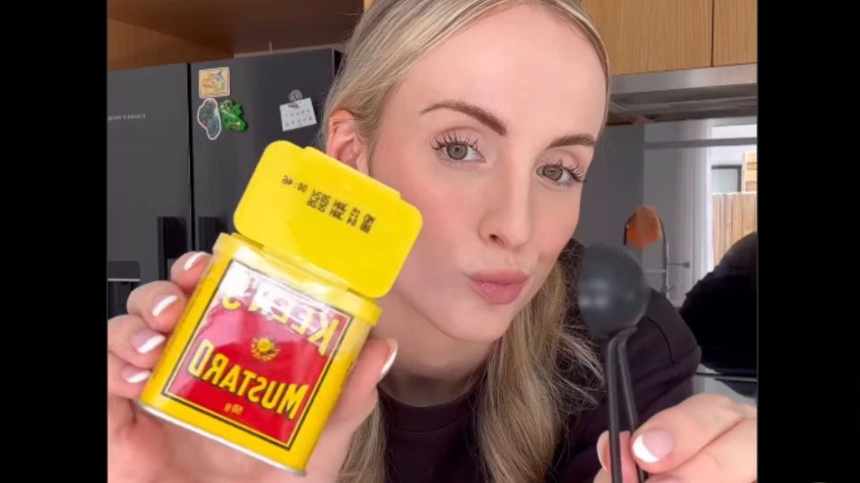 Woman holding a tin of mustard and a small black whisk.