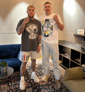 Two men standing together, one in a grey t-shirt and beige shorts, the other in a white t-shirt and light grey pants.