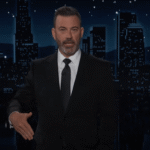 Jimmy Kimmel extending his hand in front of a cityscape at night.