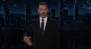 Jimmy Kimmel extending his hand in front of a cityscape at night.