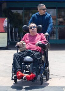Tim Curry in a wheelchair, being pushed by another person.