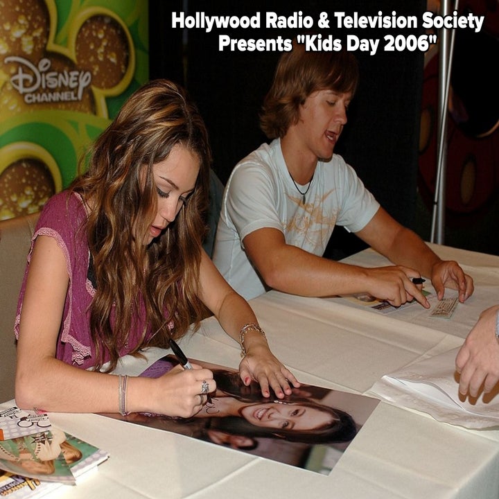 Two young people are seated at a table signing autographs at a Disney Channel event