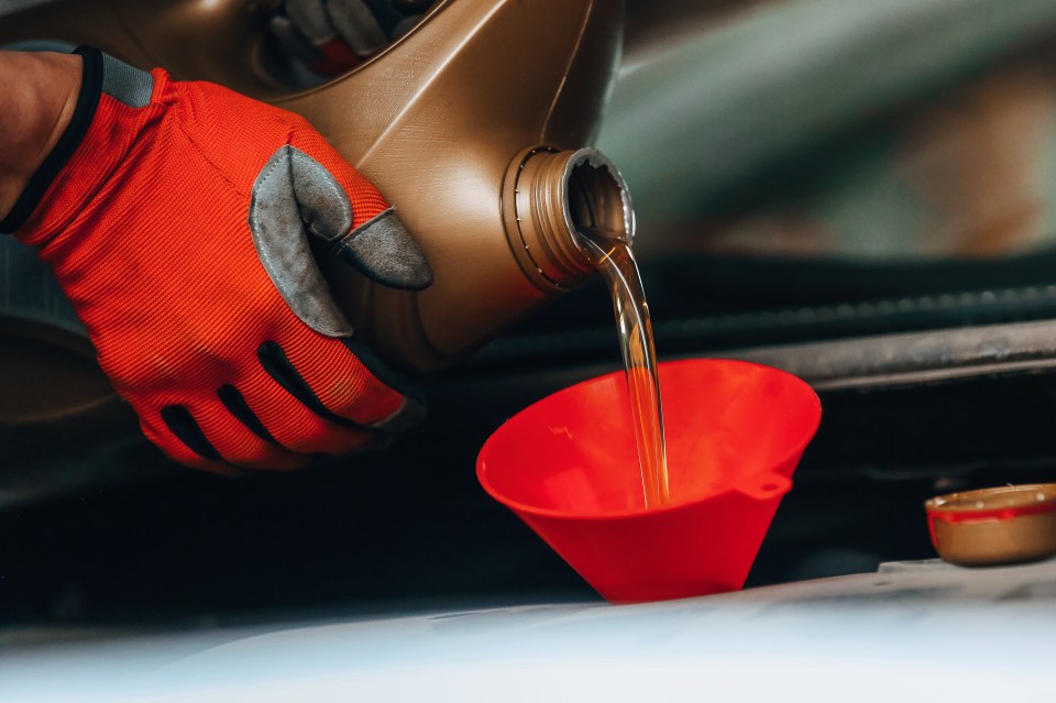 A gloved hand pouring motor oil into a red funnel.