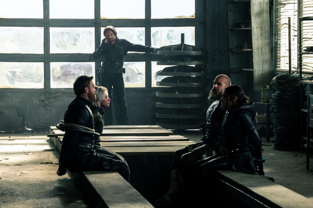 A man in a jumpsuit stands by a door while four others sit tied-up on benches in "Thunderbolts*."