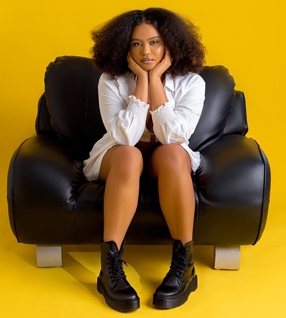 Woman with afro sitting in a black chair against a yellow background.