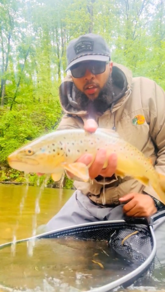Man holding a trout by a stream.