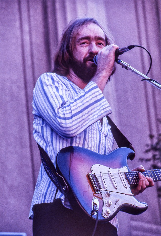 Dave Mason performing at the Greek Theater, playing guitar and singing into a microphone.