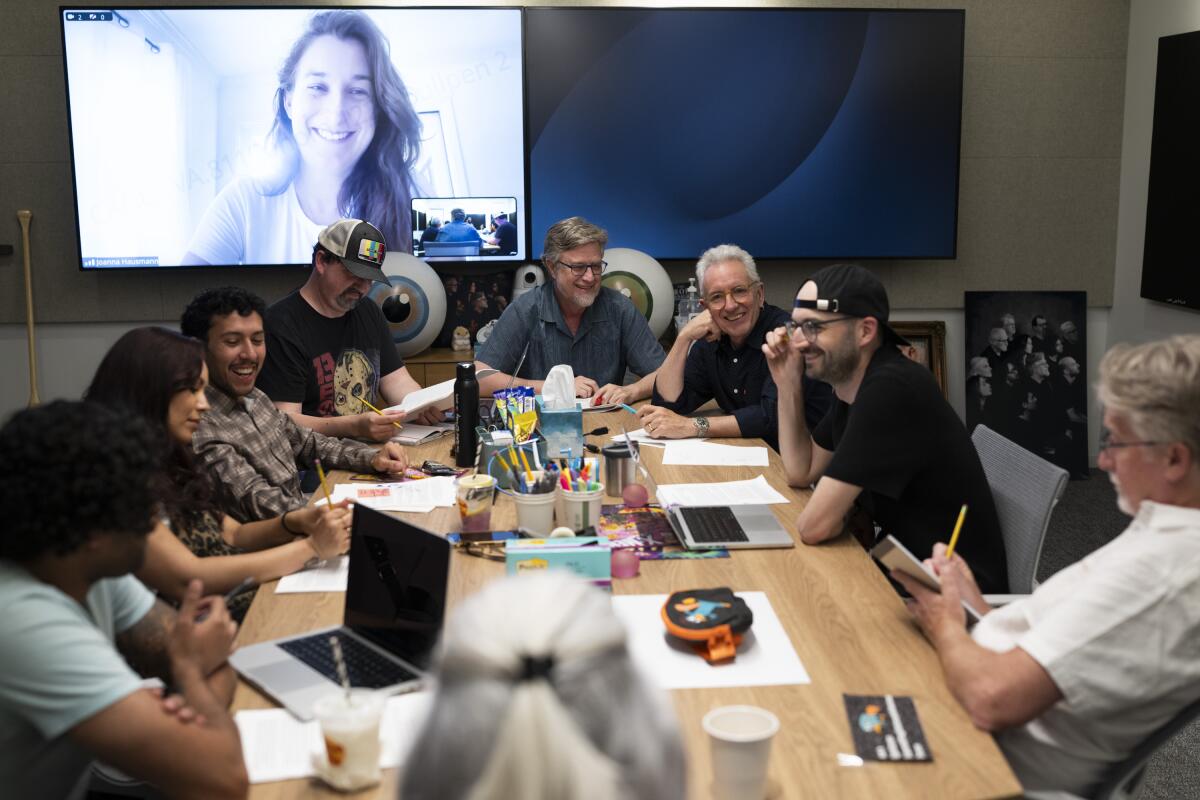 A group of people sit around a conference table and the image of a woman with long hair is seen on a screen above them.