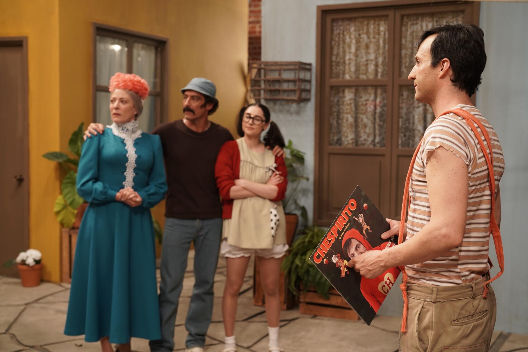 Three people stand near a doorway as another man stands opposite of them holding a Chespirito album cover.