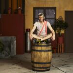 A man dressed as boy in a striped shirt, shorts and orange suspenders sits on a barrel in a courtyard.